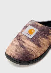 Chaussure à enfiler texturée en tissu marron et beige. Présente un patch avec le logo Carhartt et une semelle en caoutchouc noir. Conception à bout arrondi avec des détails de couture.