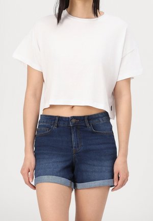 Femme portant un haut court à manches courtes blanc et un short en denim bleu foncé avec revers, sur un fond uni.