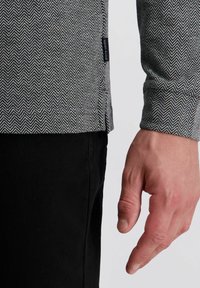 Grau und schwarz gemusterte Longsleeve-Bluse im Fischgrätmuster mitSeitenschlitz; schwarze Hosen sichtbar, die Textur und Kontrast betonen.