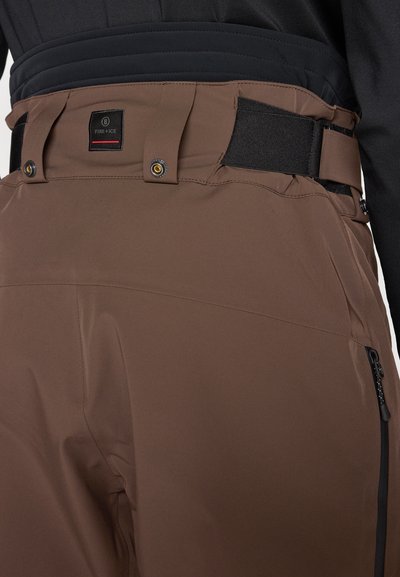 Bruna skidbyxor med svart midjeband, utrustade med justerbara remmar, fickor med dragkedja och märkespatch. Slät, vattenavvisande tyg.