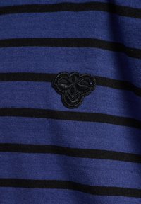 Blå randigt tyg med horisontella svarta ränder; har en svart broderad logotyp som liknar ett stiliserat insekt på vänster sida.