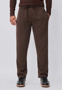 Homme portant un pantalon marron à cordon de serrage et des chaussures noires, debout les mains dans les poches devant un fond uni.