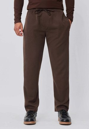 Homme portant un pantalon marron à cordon de serrage et des chaussures noires, debout les mains dans les poches devant un fond uni.