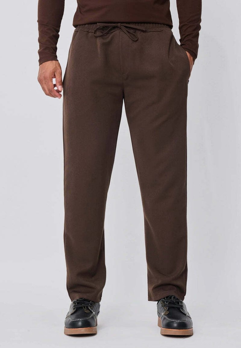 Homme portant un pantalon marron à cordon de serrage et des chaussures noires, debout les mains dans les poches devant un fond uni.