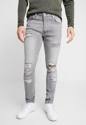 Jeans ajustados en gris con un acabado desgastado y múltiples desgarros; la tela es de denim, con desgaste en las rodillas y los muslos. Combinados con zapatillas blancas.