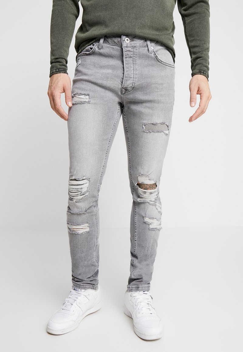 Jeans ajustados en gris con un acabado desgastado y múltiples desgarros; la tela es de denim, con desgaste en las rodillas y los muslos. Combinados con zapatillas blancas.