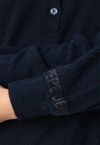 Tamsiai mėlynas megztinis su sagtimi prie apykaklės, pasižymintis tekstūruota medžiaga ir matomu logotipo detaliu ant rankovės šviesesnėje spalvoje.