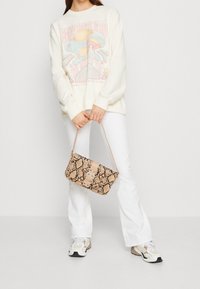 Hnědo-beige kabelka se vzorem hadí kůže a klopou s přezkou, kterou drží osoba v krémovém svetru a bílých rozšířených kalhotách.