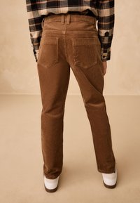 Pantalon en velours côtelé marron à coupe droite, avec deux poches arrière et une texture côtelée subtile, associé à des baskets blanches.