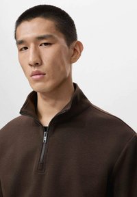 Felpa marrone con zip a un quarto, dalla texture liscia e con una zip argentata. Il colletto è rialzato e una sottostrato nero è visibile sotto.