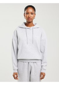 Žena okrenuta prema naprijed, nosi svijetlosivu majicu s kapuljačom i odgovarajuće trenirke, stoji na čistoj svijetloj pozadini s neutralnim izrazom lica.