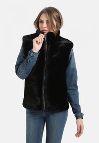 Gilet en fausse fourrure noire avec col montant et fermeture zip, superposé sur une veste en denim. Présente une texture douce et une coupe ajustée.