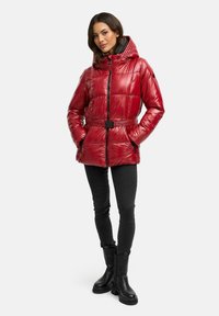 Rote Steppjacke mit Kapuze, mit gestepptem Design, Reißverschluss vorne, elastischem Bund und kontrastierendem schwarzen Gürtel. Kombiniert mit schwarzen Hosen und Stiefeln.