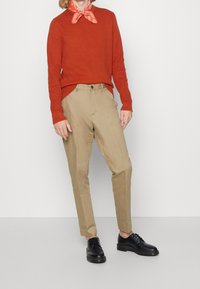 Personne portant un pull orange, un pantalon beige, un bandana rouge autour du cou et des chaussures noires à lacets, debout devant un fond blanc uni.