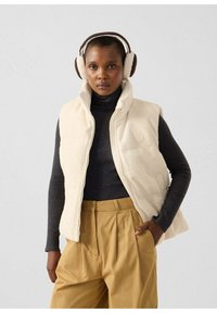 Gilet in pile color crema con collo alto e zip, abbinato a un dolcevita a coste nero e pantaloni kaki a vita alta. Completano il look le cuffie marroni.