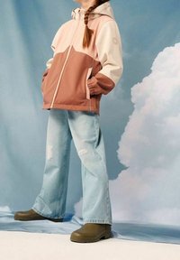 Farbig blockierte Kapuzenjacke in Pink, Braun und Creme, kombiniert mit hellblauen, ausgewaschenen Jeans und dunkelgrünen Knöchelstiefeln.
