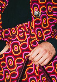 Abito colorato con motivo a grandi forme ovali intrecciate in rosso, arancione, rosa e marrone. Dettaglio visibile di bottoni e cintura.