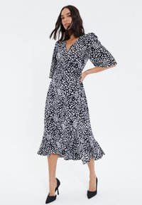 Vestido negro con lunares blancos, escote en V, mangas abullonadas y dobladillo con volantes. Combina con tacones altos negros. Textura de tela suave.