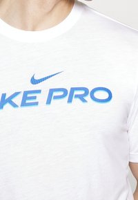Vit Nike t-shirt med blå "NIKE PRO" text och randiga detaljer. Mjuk tyg, rund hals, kort ärm, och inga ytterligare mönster.