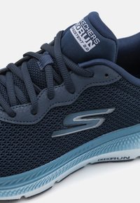 Pantofii sport de culoare bleumarin cu plasă în partea superioară, sistem de închidere cu șirete, detalii texturate și talpă intermediară de culoare albastru deschis; au logo-ul Skechers pe lateral.