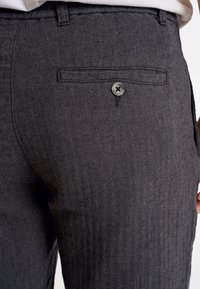 Pantalons en chevrons gris foncé, avec une texture lisse, dotés d'une poche arrière avec un fermeture à bouton. Aucun motif ou accent visible.