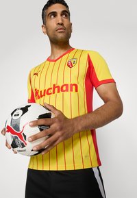 Un uomo in una maglia da calcio a righe gialle e rosse tiene in mano un pallone da calcio bianco e nero, guardando con sicurezza verso l'alto su uno sfondo neutro.