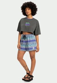 Pilkas sutrumpintas marškinėliai su violetiniais akcentais ir grafika, derinami su mėlynomis raštuotomis maudymosi šortais, turinčiais raištelį ir logotipo pleistrą.