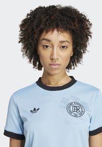Lys blå sportstrøye med svarte detaljer, ribbet stoff, korte ermer og et sirkulært logo på brystet, med et vintage design.