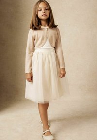 Cardigan scurt de culoare roz pal, top alb ajustat, fustă din tulle stratificată în nuanță de crem. Textură moale, decolteu rotund și pantofi cu vârf închis de culoare deschisă.