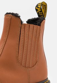 Brązowy skórzany botek za kostkę z miękką czarną futrzaną podszewką, elastycznymi bocznymi panelami i żółtą pętlą do zakupu. Posiada kontrastowe przeszycia wokół krawędzi.