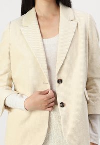 Beige korduroy blejzr s klopovým límcem, se dvěma tmavými knoflíky a oversized střihem. Dlouhé rukávy s detaily na manžetách. Texturovaná tkanina.