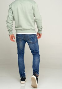 Hombre caminando alejándose, vistiendo un sudadera verde claro, jeans azules desgastados, zapatillas negras y un reloj en la muñeca izquierda, sobre fondo blanco.