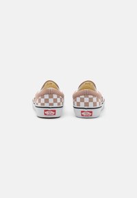 Zapatillas de lona rosa sin cordones con un patrón de tablero de ajedrez blanco en los laterales. Cuentan con una suela blanca con una franja negra y una etiqueta de logo roja.