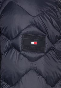 Marineblå pufferjakke med en kviltet design, der har et sort rektangulært logo-patch med røde og hvide accenter. Glat, blank tekstur.
