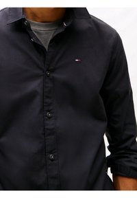 Camisa de botones azul marino oscuro con cuello. Presenta un pequeño logo en el pecho, botones negros y mangas remangadas para un estilo relajado.