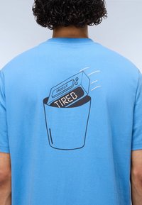 Μπλε βαμβακερό μπλουζάκι με μαύρο σχέδιο ψηφιακού ξυπνητηριού που φέρει την ένδειξη «SNOOZE» και φαίνεται να ξεπροβάλλει από μια τσέπη, με την ένδειξη «TIRED».