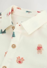 Weißes Hemd mit verspielten Druckmustern, die Lebkuchenmänner, Rentiere und Weihnachtsbäume zeigen. Holzknöpfe am Kragen.