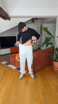 Junge Person mit schwarzer Kappe und schwarzem Shirt hebt das Shirt an, während sie in einem hellen Wohnzimmer mit Couch, Fernseher und großer Pflanze weiße Jogginghose richtet.
