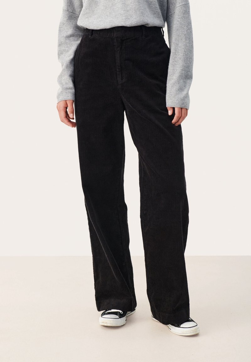 Personne portant un pantalon large en velours côtelé noir, un pull gris et des baskets noir et blanc, debout devant un fond uni.