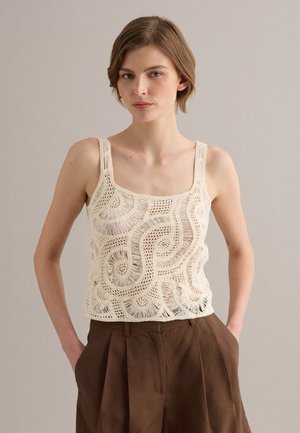 Giovane donna con capelli corti che indossa un top all’uncinetto color crema e pantaloni marroni a vita alta con pieghe, su uno sfondo neutro.