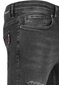 Jeans de mezclilla negra con corte ajustado, que presentan detalles desgastados en la rodilla, un pequeño bolsillo con un acento rojo y costuras horizontales en la cinturilla.