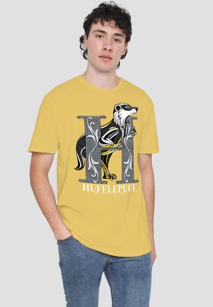 Man wearing yellow Hufflepuff T-shirt with large decorative "H" and badger graphic, paired with blue jeans, standing against plain background.