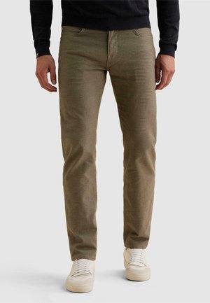 Homme portant un pantalon vert olive et des baskets blanches, debout devant un fond uni, les mains relâchées le long du corps.