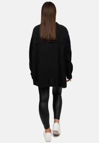 Camicia nera oversize in tessuto testurizzato con maniche lunghe, caratterizzata da un orlo asimmetrico, abbinata a leggings neri e sneakers bianche.