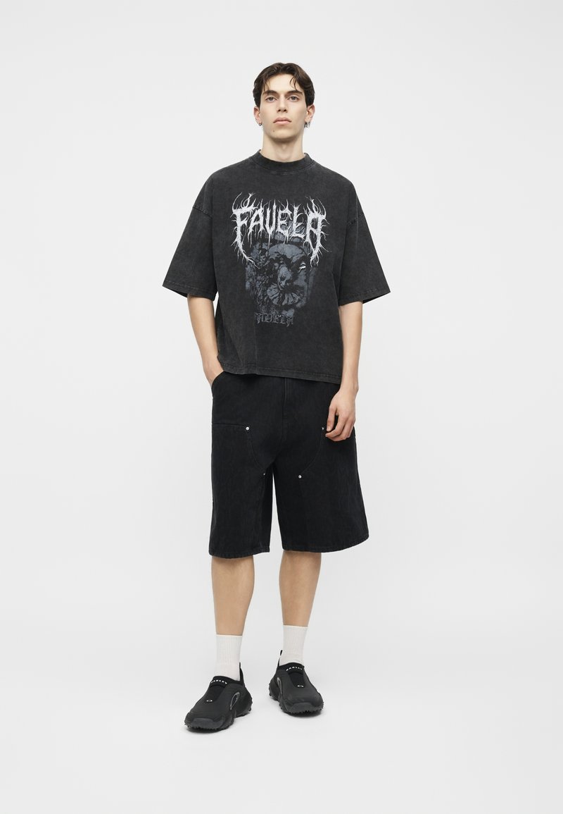 Junger Mann steht vor weißem Hintergrund, trägt ein übergroßes schwarzes T-Shirt mit Grafik, schwarze knielange Shorts, weiße Socken und schwarze Slip-On-Schuhe.