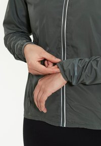 Donkergrijze jas van lichtgewicht, glad materiaal met reflecterende accenten en ritszakken. Elastische manchetten worden aangepast.