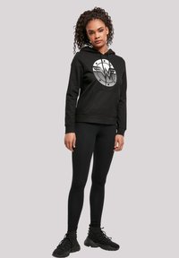 Black hoodie with a Wonder Woman logo, front pocket, and drawstring hood. Paired with black leggings and sporty black shoes.