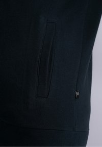 Ciemnoniebieski sweatshirt z boczną kieszenią i żebrowaną teksturą. Zawiera małą metkę marki przy szwie w celu identyfikacji.