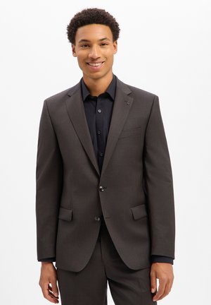 Man wearing a dark brown suit and black shirt, standing and smiling against a plain white background.