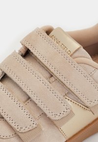 Tuvumā iemūžināta bežā zamšā pagatavota sporta apava ar trim velcro siksniņām un zelta akcentu uz mēles, kurā redzama zīmola nosaukums.
